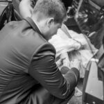 Der Bräutigam verschließt den Schuh der Braut, während sie vergnüglich im schwarzen Cabrio sitzt und den Bräutigam anlächelt