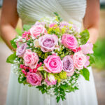Braut hält einen romantischen Hochzeitsstrauß aus rosa und lila Rosen, verziert mit Schleierkraut und grünem Blattwerk.